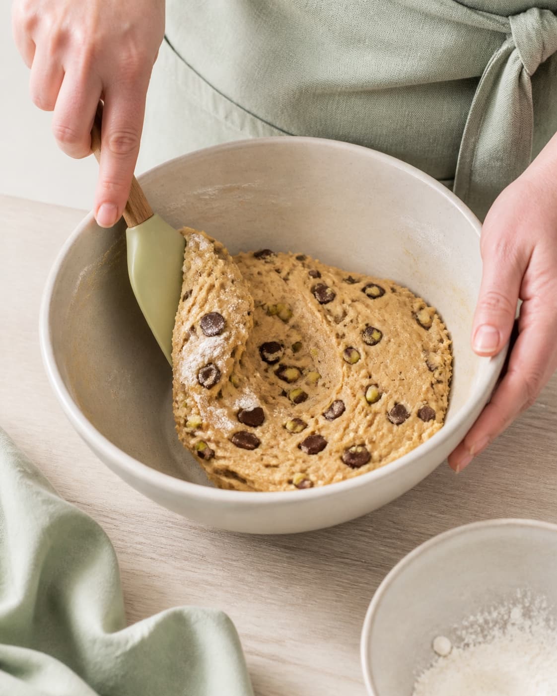 To hænder folder de sidste fine melstriber, grøn pistacie og mørke chokoladestykker skånsomt i en glat tæt cookie-dej