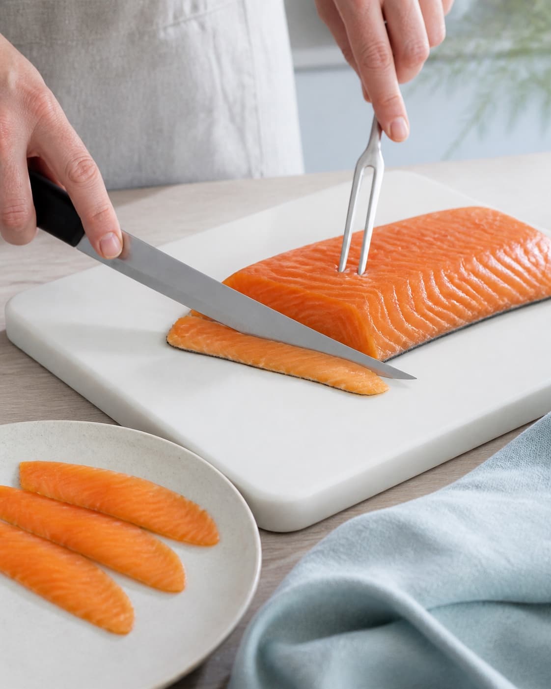 En lang kniv skærer en tynd skrå skive af fast gravad laks væk fra det synlige skind, mens en ren gaffel holder sikkert bag kniven