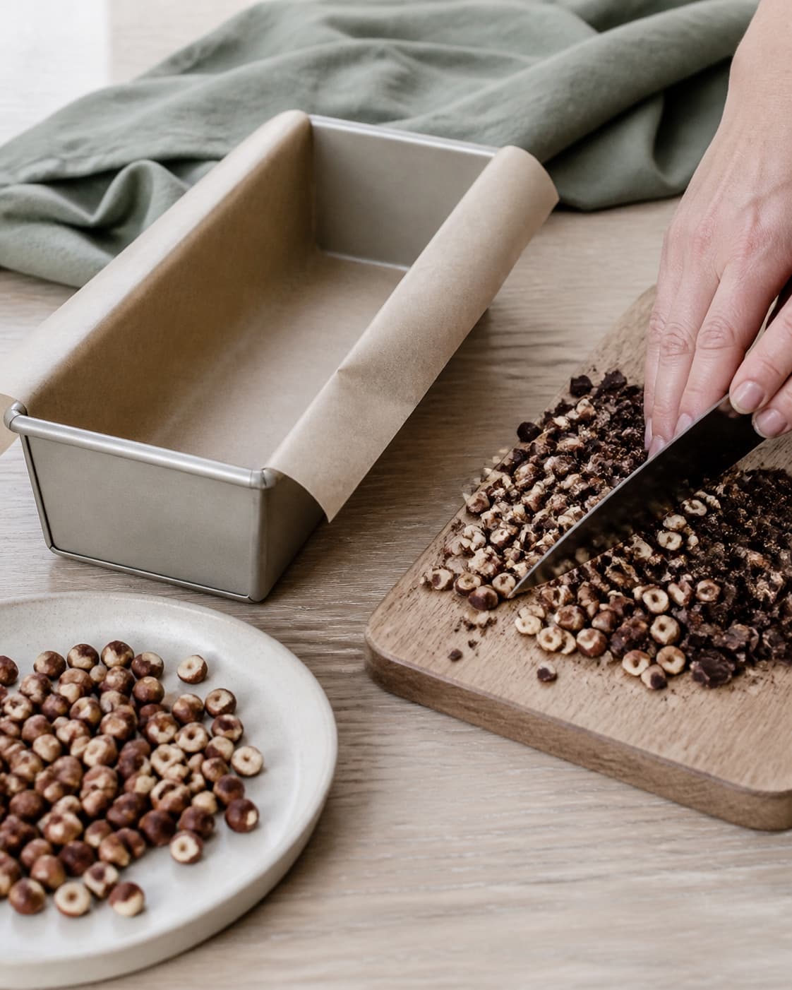 En hånd hakker afkølede ristede hasselnødder og mørk chokolade ved en brødform beklædt på langs med bagepapir