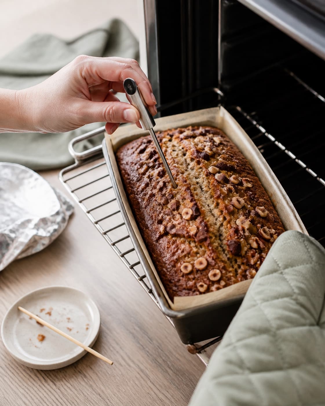 En hånd måler midten af det gyldne revnede bananbrød med kernetermometer, mens en ovnhandske beskytter ved formen