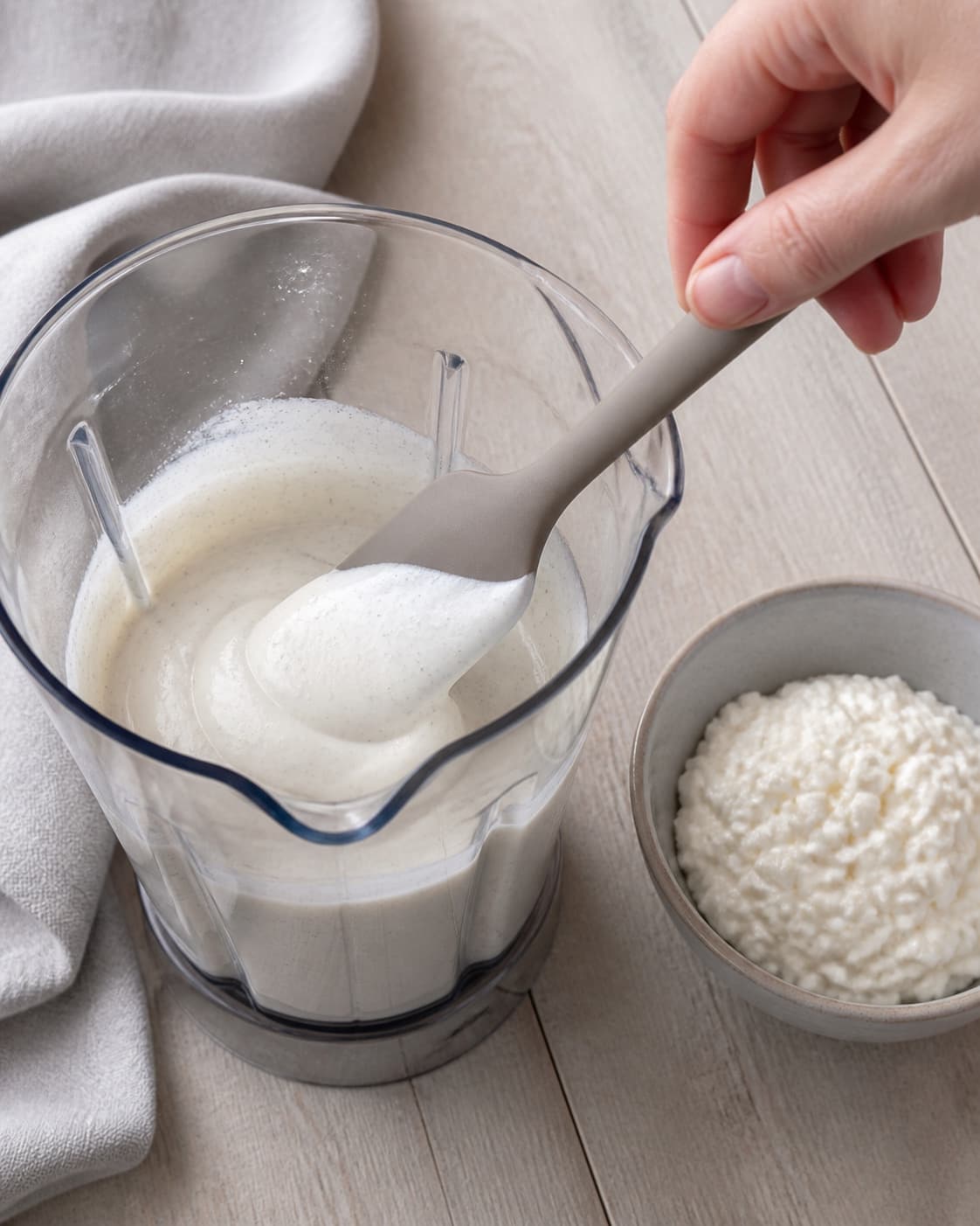 En hånd viser den tykke glatte vaniljebase af skyr og hytteost i blenderen ved en separat skål med reserveret hytteost