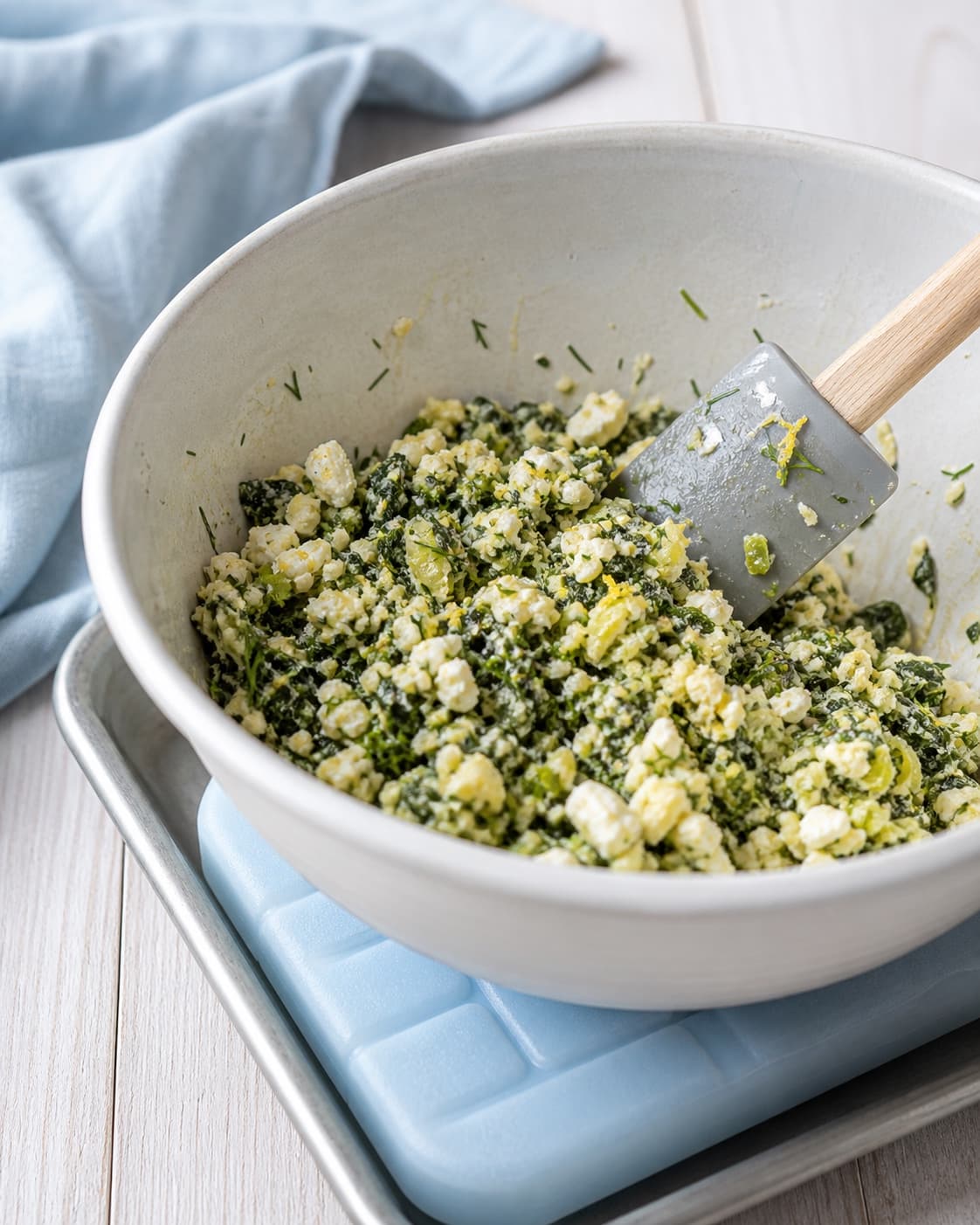 En dejskraber vender det afkølede spinatfyld med feta, dild og citronskal i en skål over en kold blå køleblok