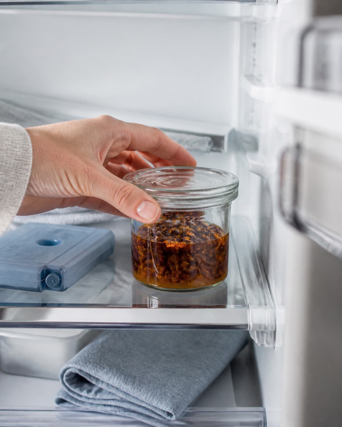 En hånd vender det lukkede glas med rosiner, rom, appelsinsaft og skal på en ren køleskabshylde ved siden af en køleblok