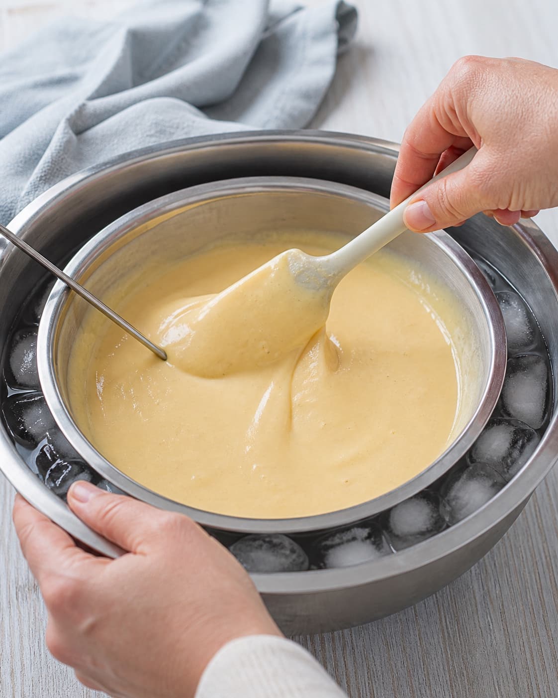 En dejskraber rører den silede blanke æggecreme i en lav metalbeholder omgivet af isvand, mens temperaturproben bliver i cremen
