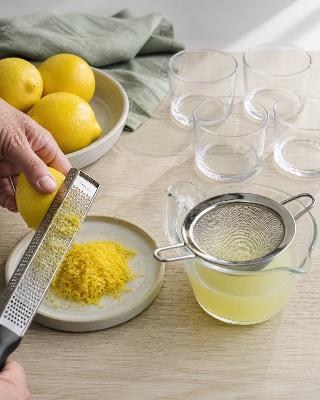 En hånd river kun den gule skal af en vasket citron ved siden af siet kernefri citronsaft og fire rene tomme glas