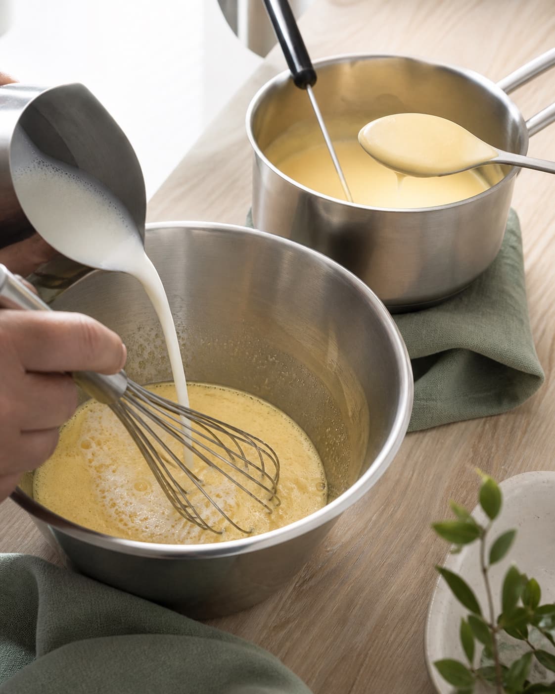 Varm mælk og fløde hældes langsomt i lyse æggeblommer under piskning, mens den glatte legerede creme kontrolleres i gryden bagved