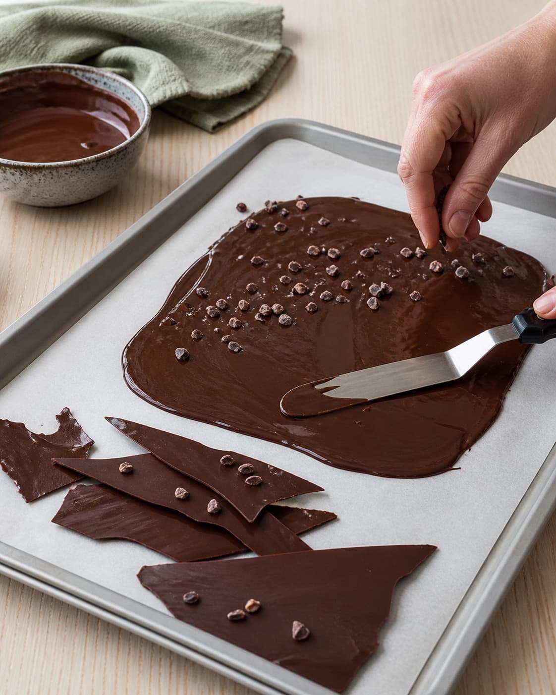 Mørk chokolade stryges papirtyndt på bagepapir og drysses med kakaonibs ved siden af helt faste sprøde chokoladeskår