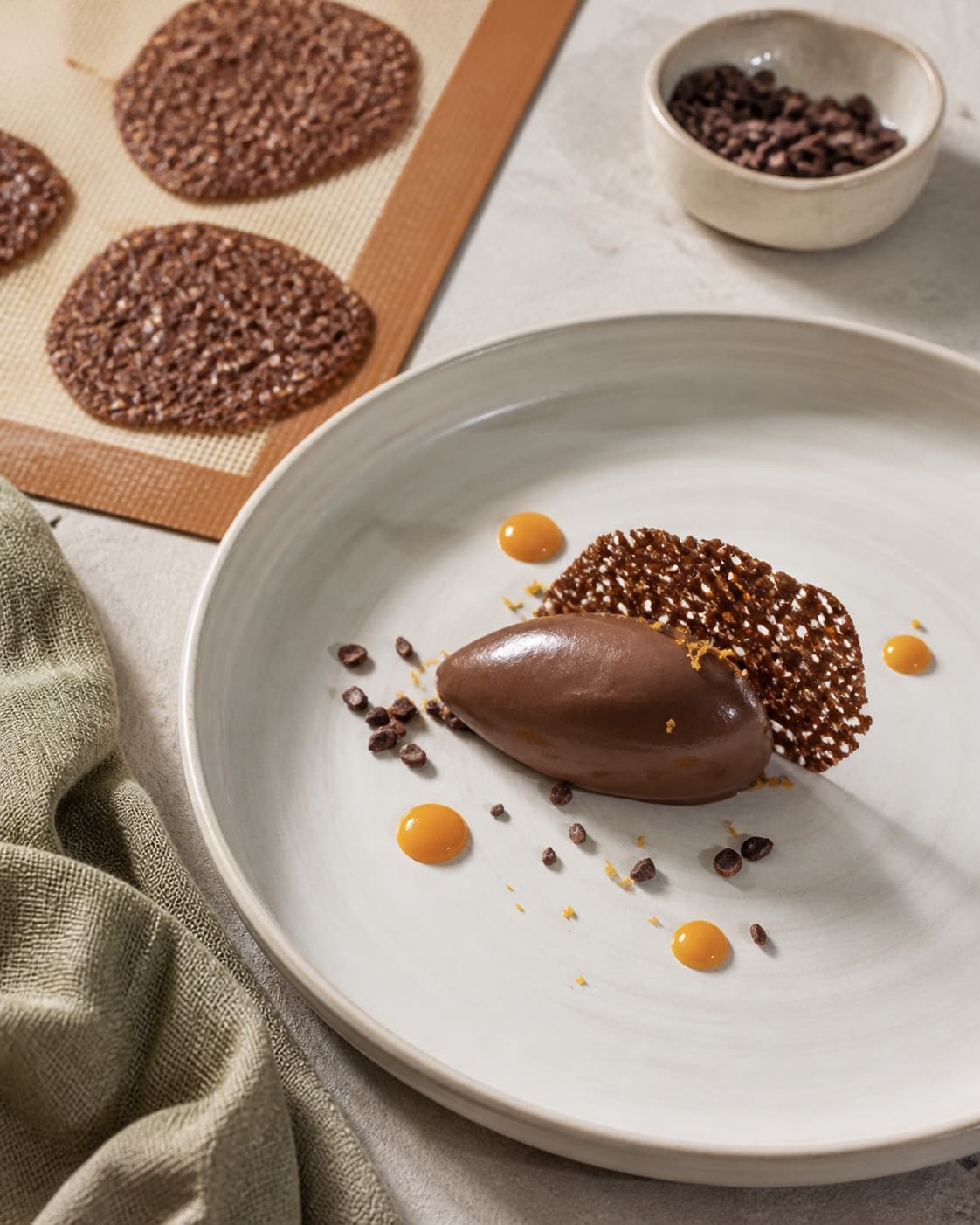 En kold glat chokolademousse-quenelle står fast med sprød ultratynd kakaotuile, kakaonibs og kontrollerede appelsinprikker