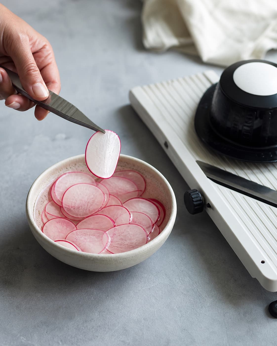 Tynde sprøde radiseskiver med rosa kant ligger i klar kold syltelage ved en mandolin med sikker håndbeskytter