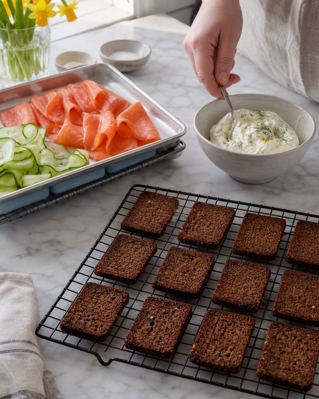 Tolv ristede rugbrødsrektangler køler enkeltvis på rist, adskilt fra kold dillcreme, røget laks og agurk på kølebakke
