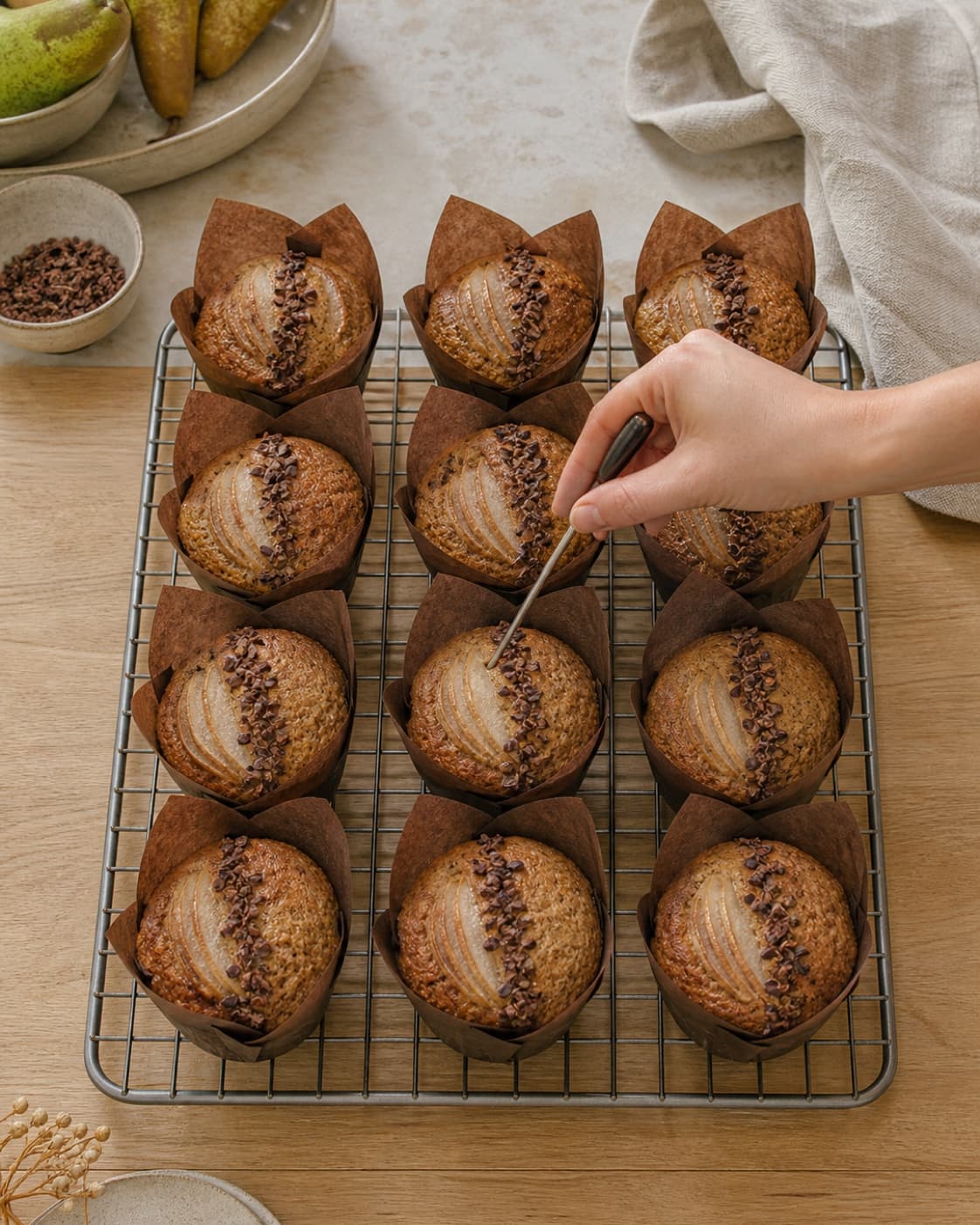 Præcis tolv høje gyldne dadelmuffins køler i tre kolonner med fire, mens en stor muffin måles i centrum ved siden af pære og kakaonibs