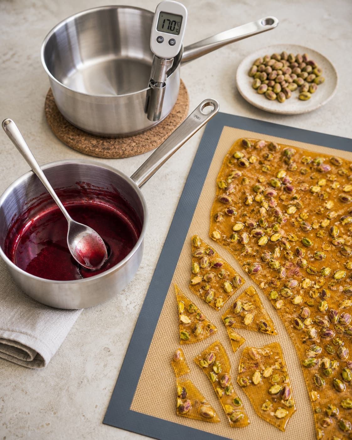 Blank mørkerød kirsebærreduktion står ved tynd lys ravfarvet pistaciebrittle i sprøde flager, mens karameltemperaturen viser 170 °C