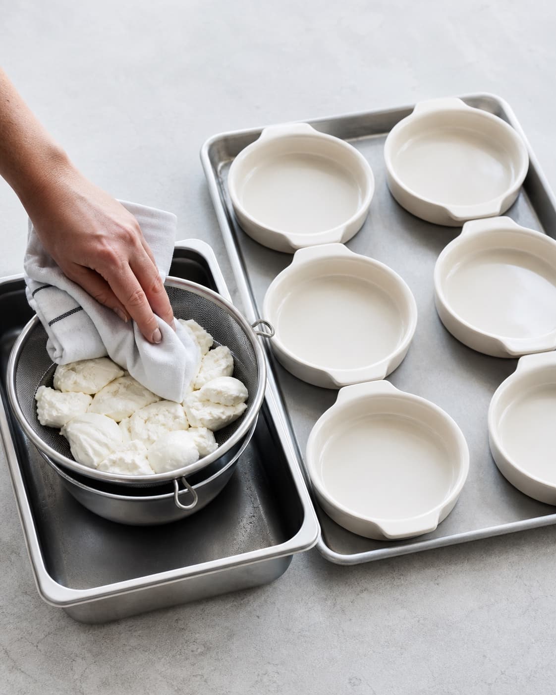 Mat mozzarella drænes og duppes i en si ved siden af præcis seks tomme cremefarvede varmefaste portionsfade på en bageplade