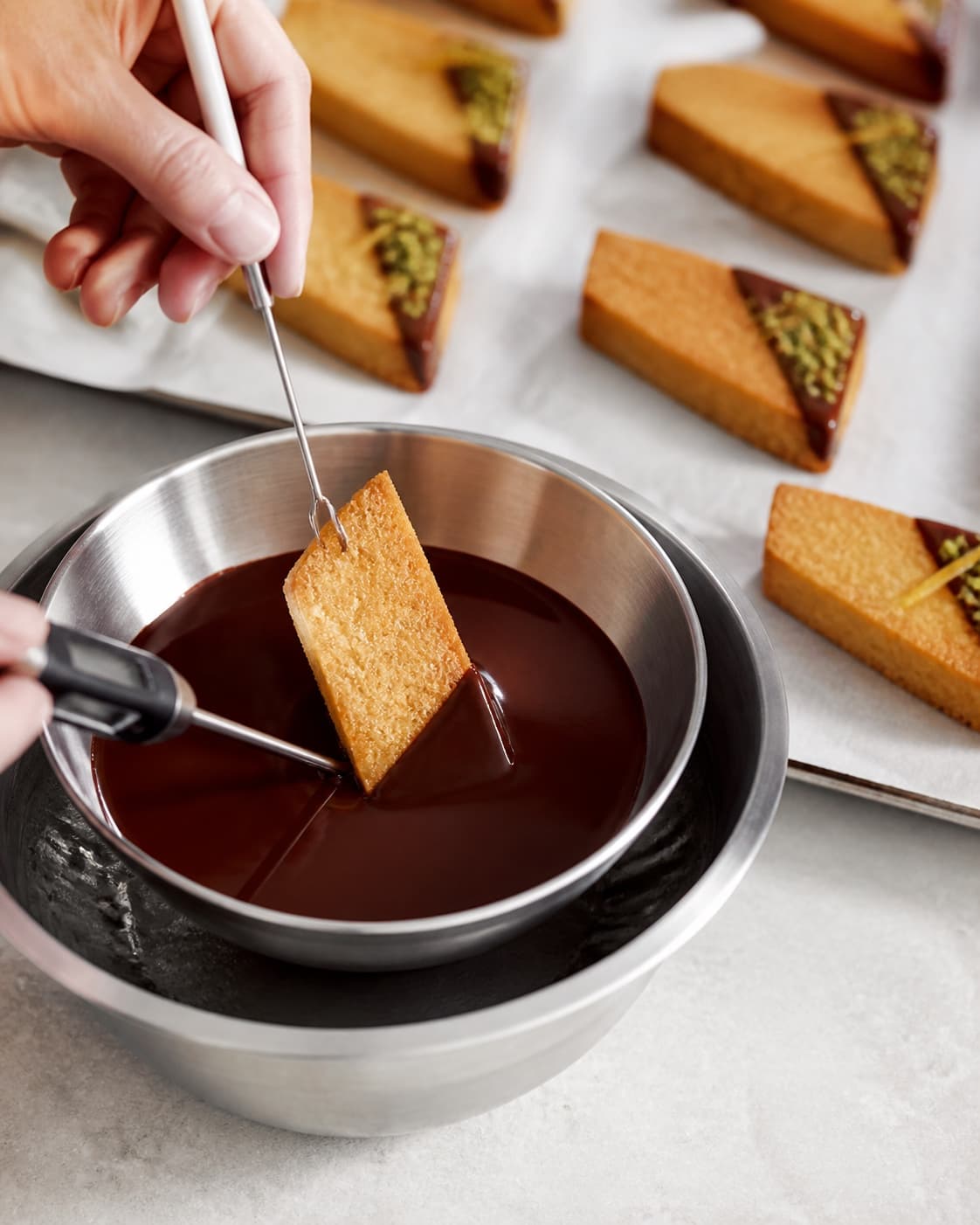 Et diagonalt hjørne af hver mazarinpetitfour dyppes i blank tempereret mørk chokolade og drysses med pistacie og citron