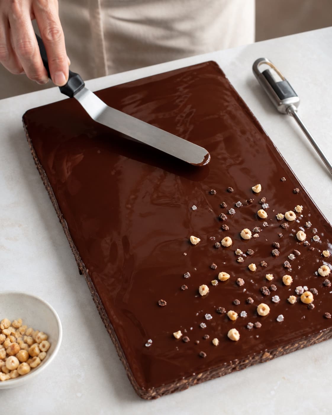 Blank mørk chokolade glattes i et tyndt lag over pladen og drysses sparsomt med nøddeknas, kakaonibs og salt