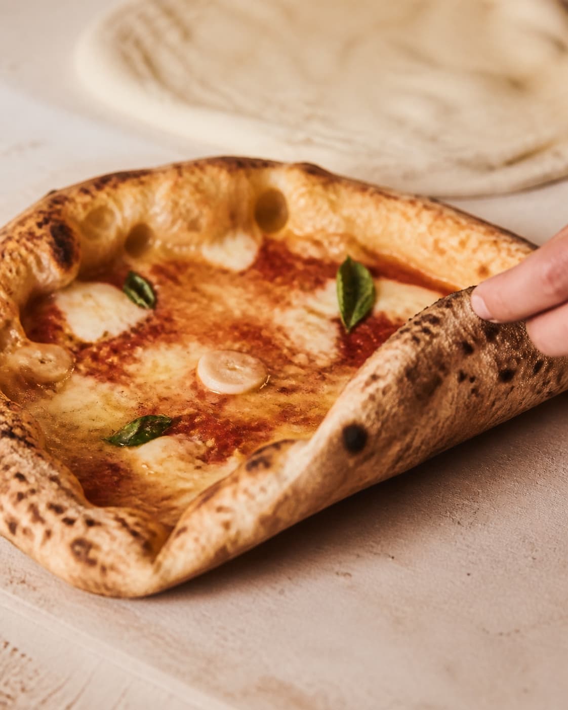 En bagt pizzakant løftes og viser tør brunplettet underside, høj blisteret rand og fuldt bagt luftig krumme