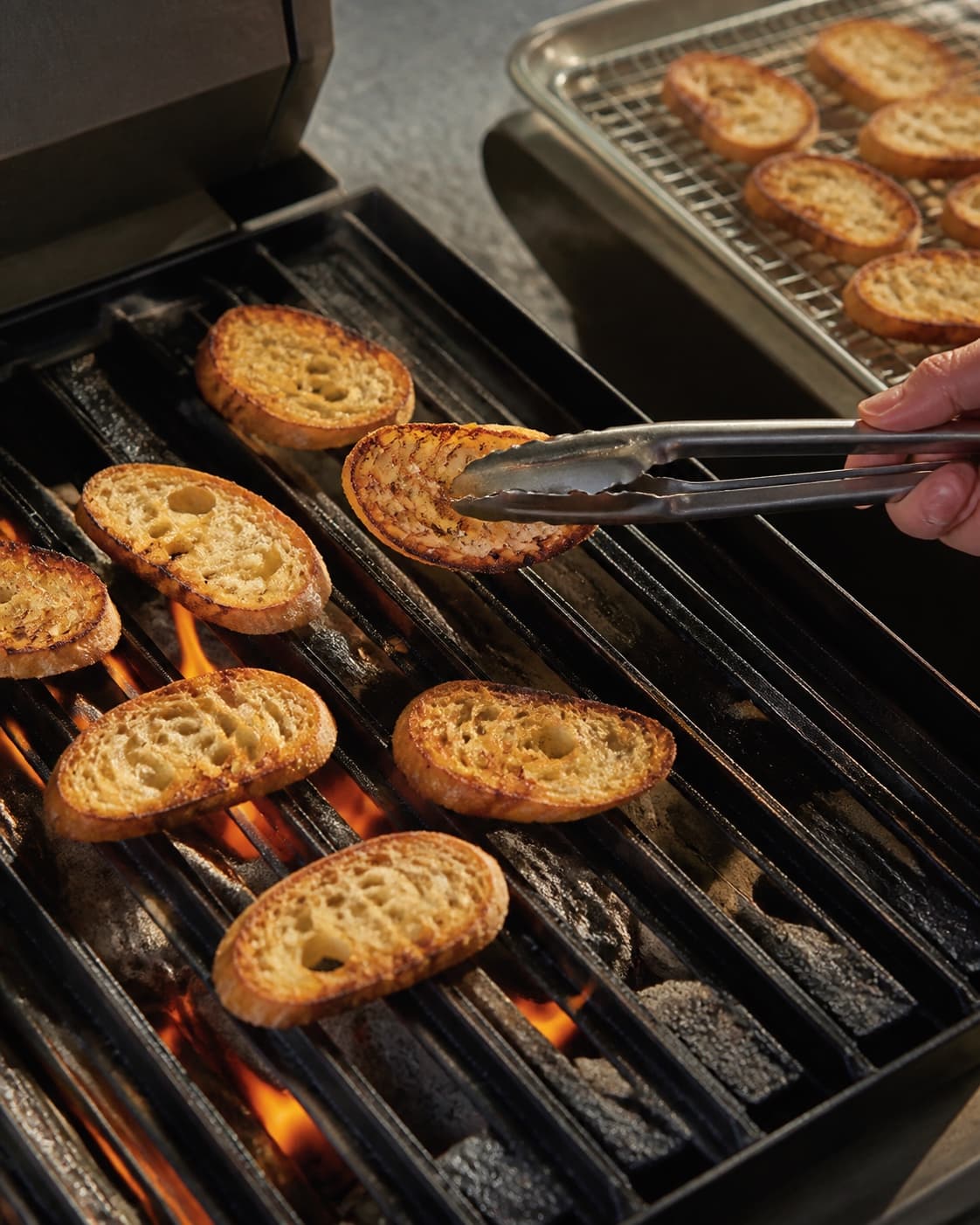 En baguetteskive løftes fra grillen med dybt gylden tør krumme, mens færdige crostini køler på rist