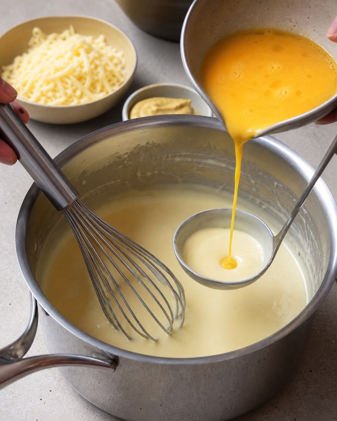 Tempereret æg hældes langsomt i glat afkølet ostesauce med sennep og revet havarti klar ved siden af