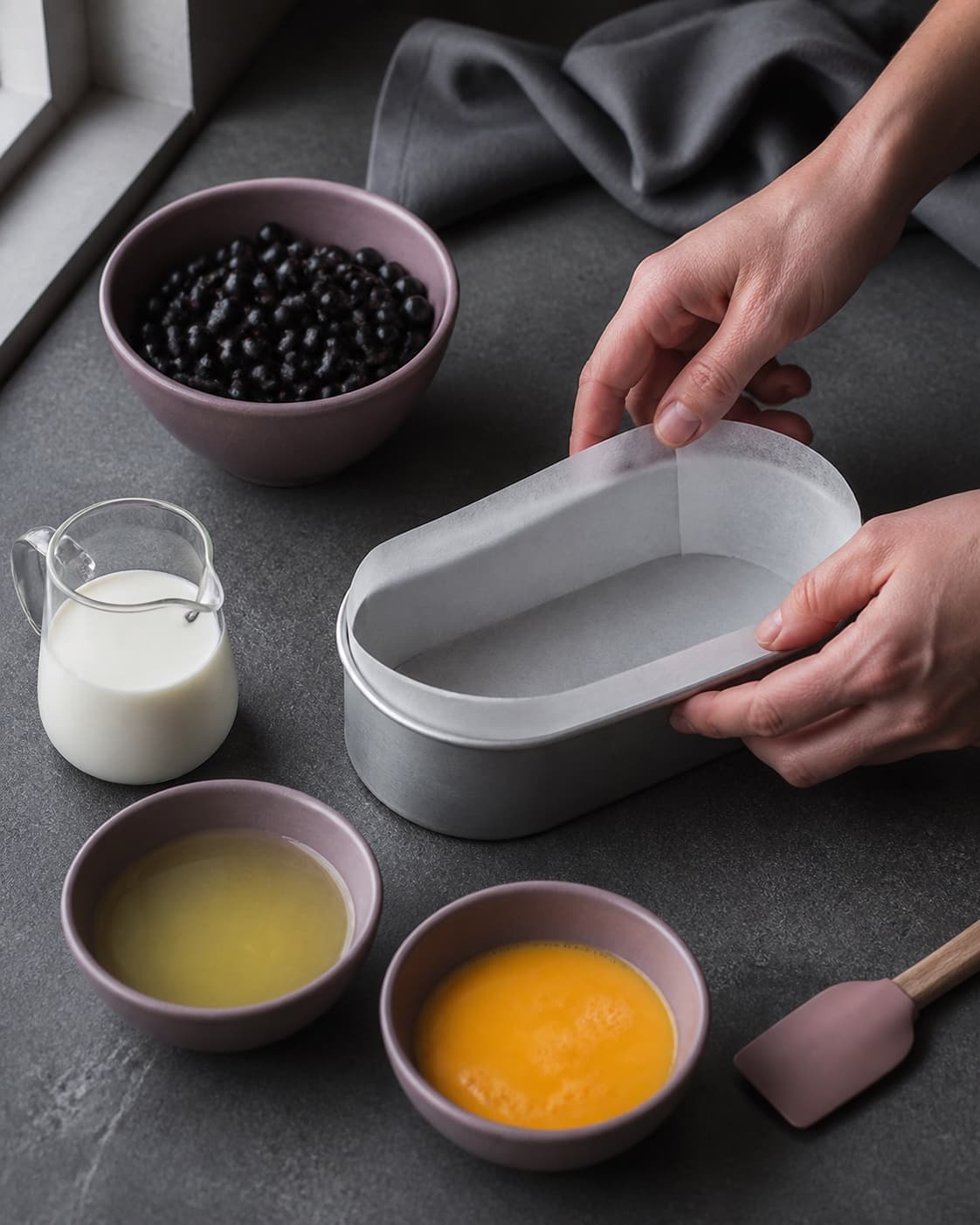 Hænder beklæder en oval kageform med bagepapir ved siden af tørre solbær og kolde pasteuriserede ægprodukter