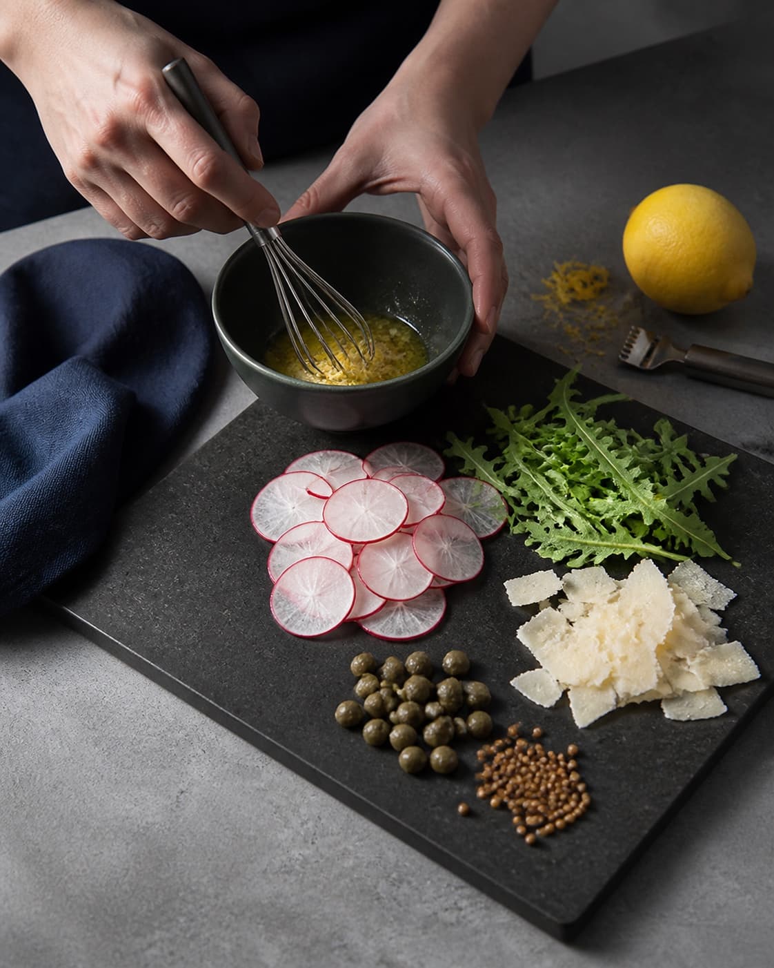 Citronolie piskes ved siden af radiseskiver, rucola, parmesan, kapers og syltede sennepsfrø på rent bræt