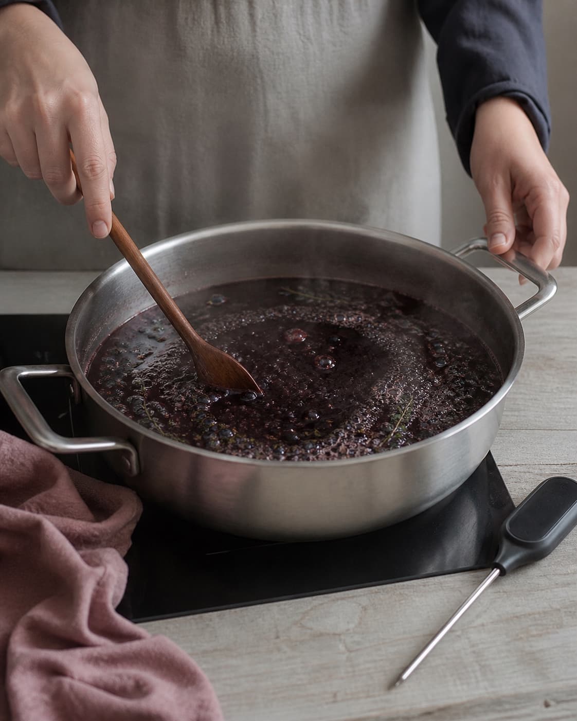 To hænder rører den endnu tynde balsamico- og druebase med synlig timian og enebær under kontrolleret opkog