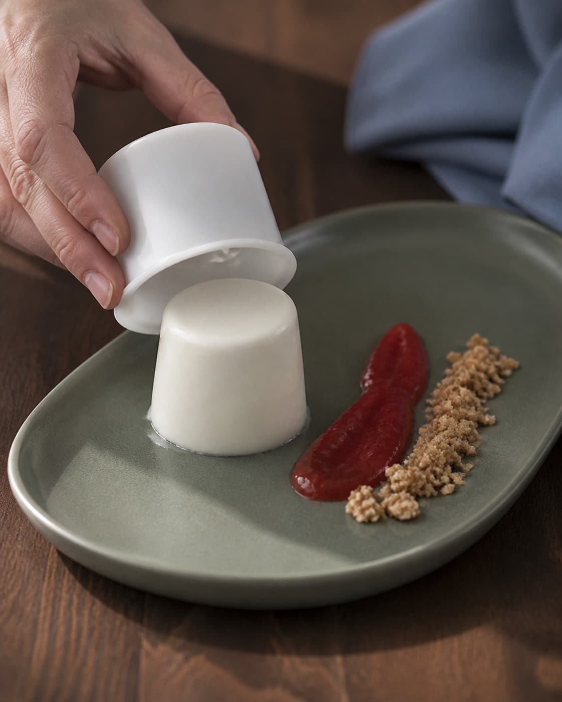 To hænder løsner en kold panna cotta fra silikoneform på salviegrønt fad og lægger kompot og crumble separat
