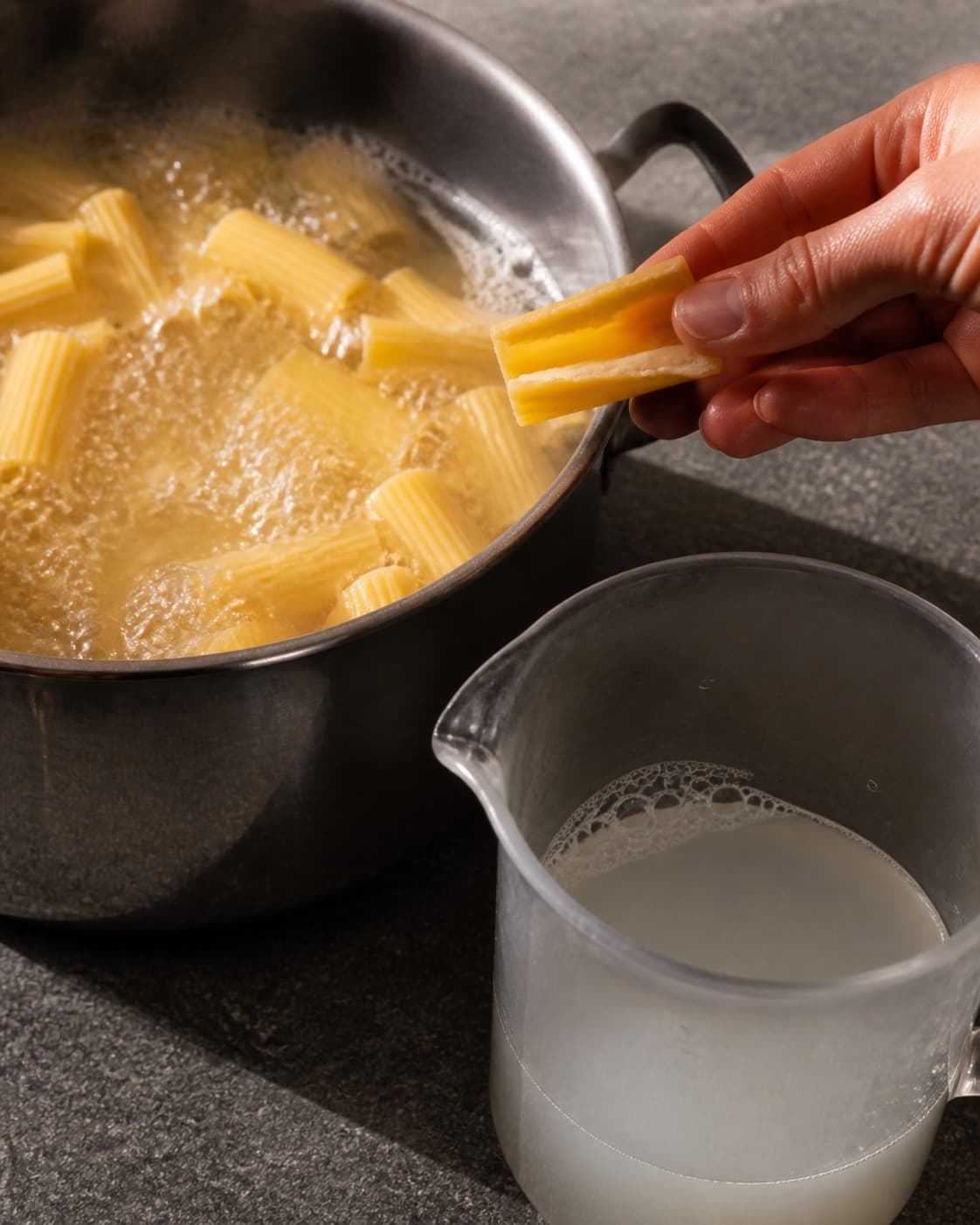 To hænder løfter én rigatoni fra saltet kogevand og bryder den for at kontrollere en tynd fast kerne