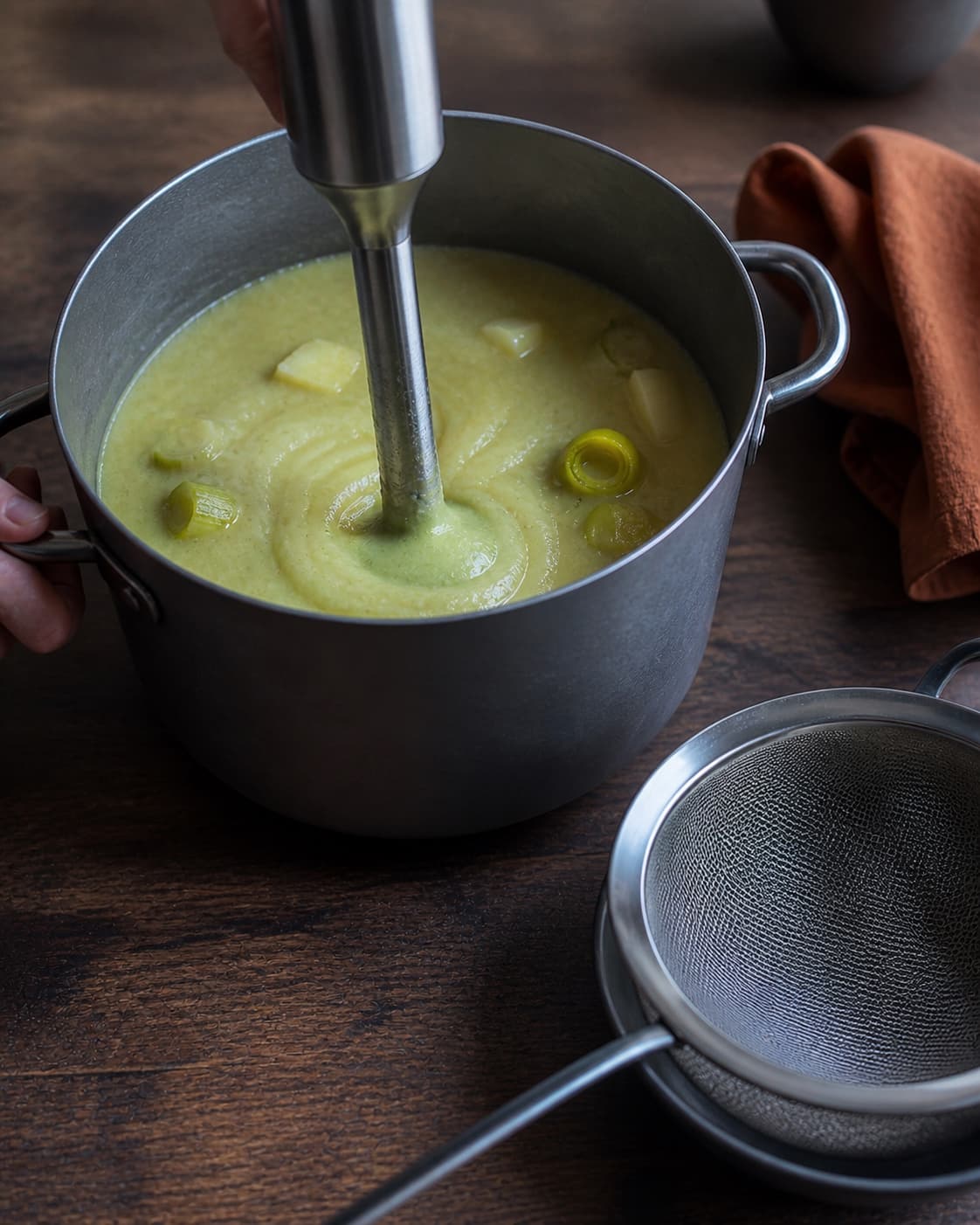 To hænder stavblender gennemkogte porrer og rodfrugter i høj gryde til helt glat palegrøn suppe