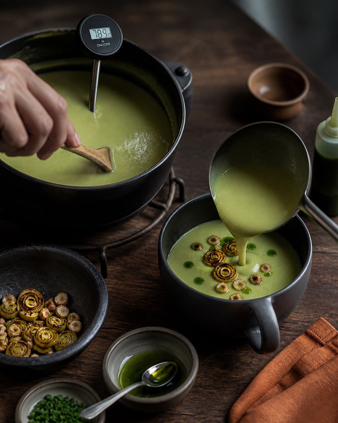 To hænder rører fløde og citron i varm glat porresuppe og anretter brunede ringe, nød og purløgsolie sent