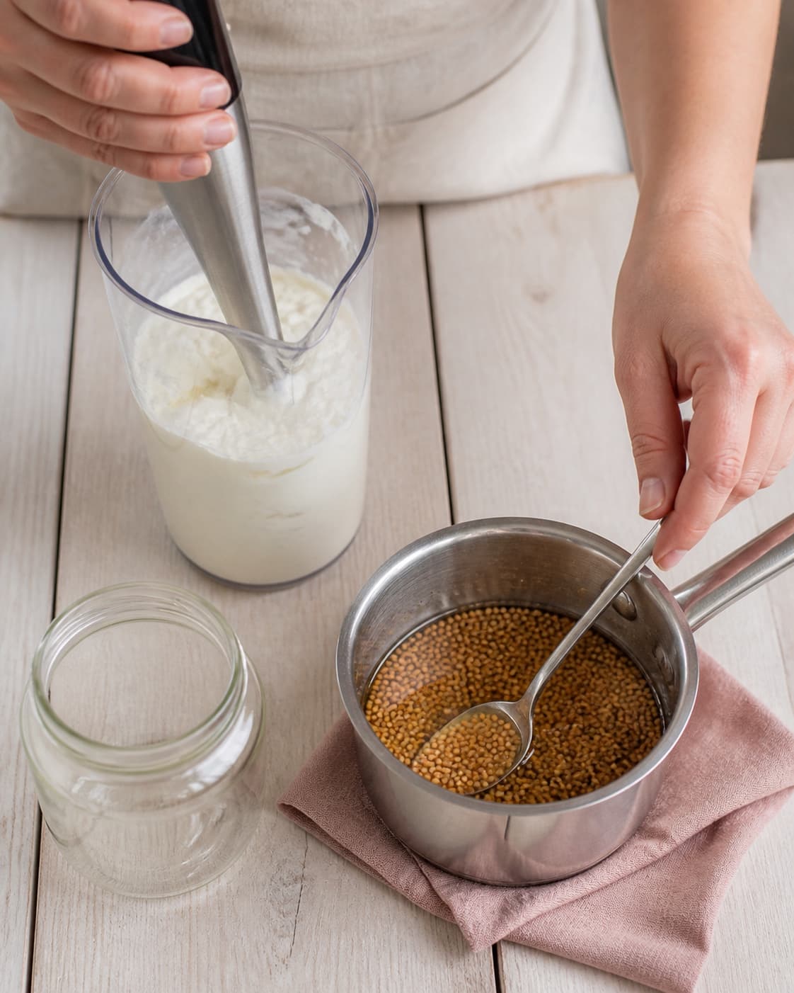 To hænder stavblender tyk feta-yoghurtcreme i høj kande og rører blanke gule sennepsfrø i varm klar lage