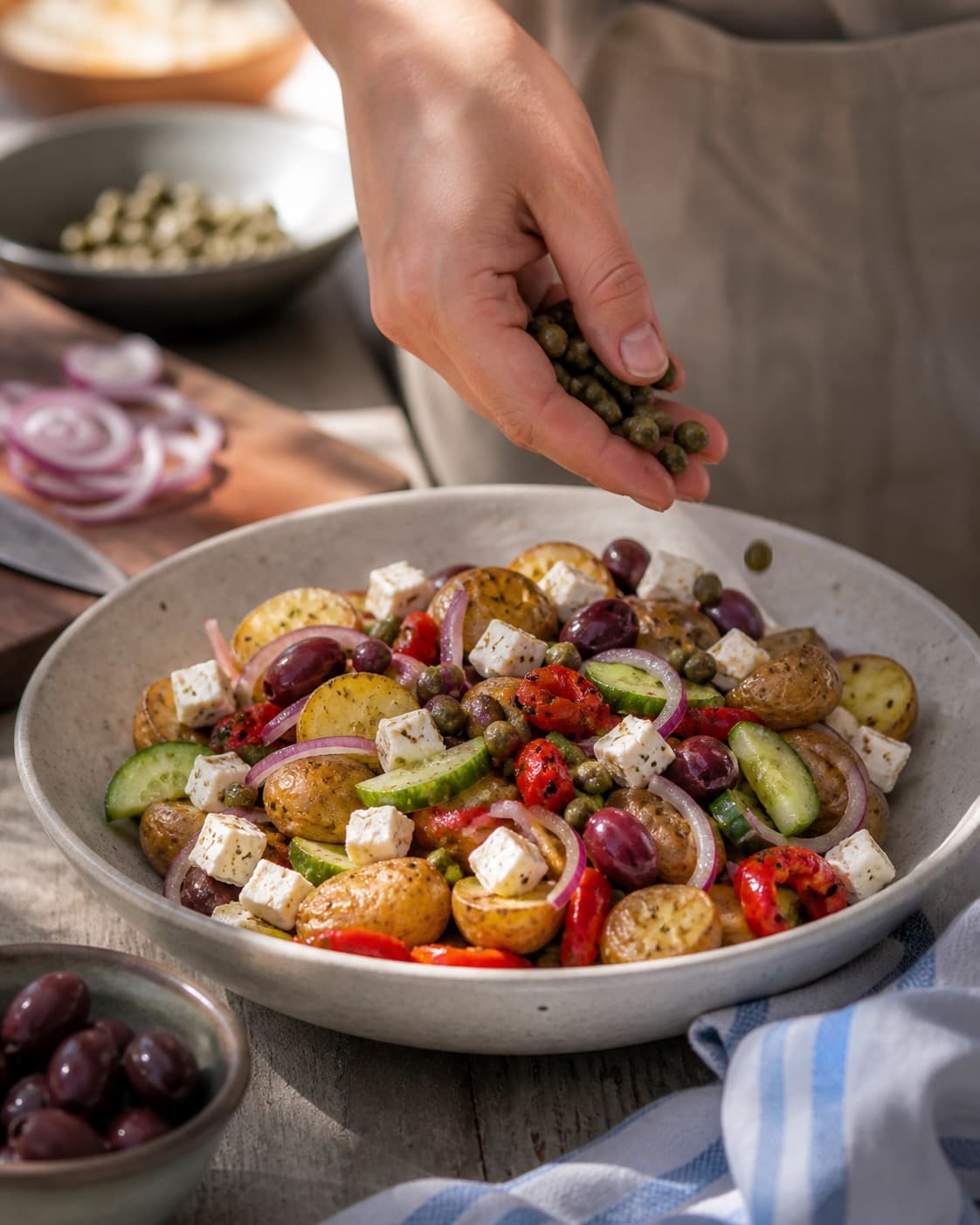 den græske kartoffelsalat anrettes luftigt med feta, kapers og rødløg under tilberedningen af græsk kartoffelsalat med feta, peberfrugt og kapers