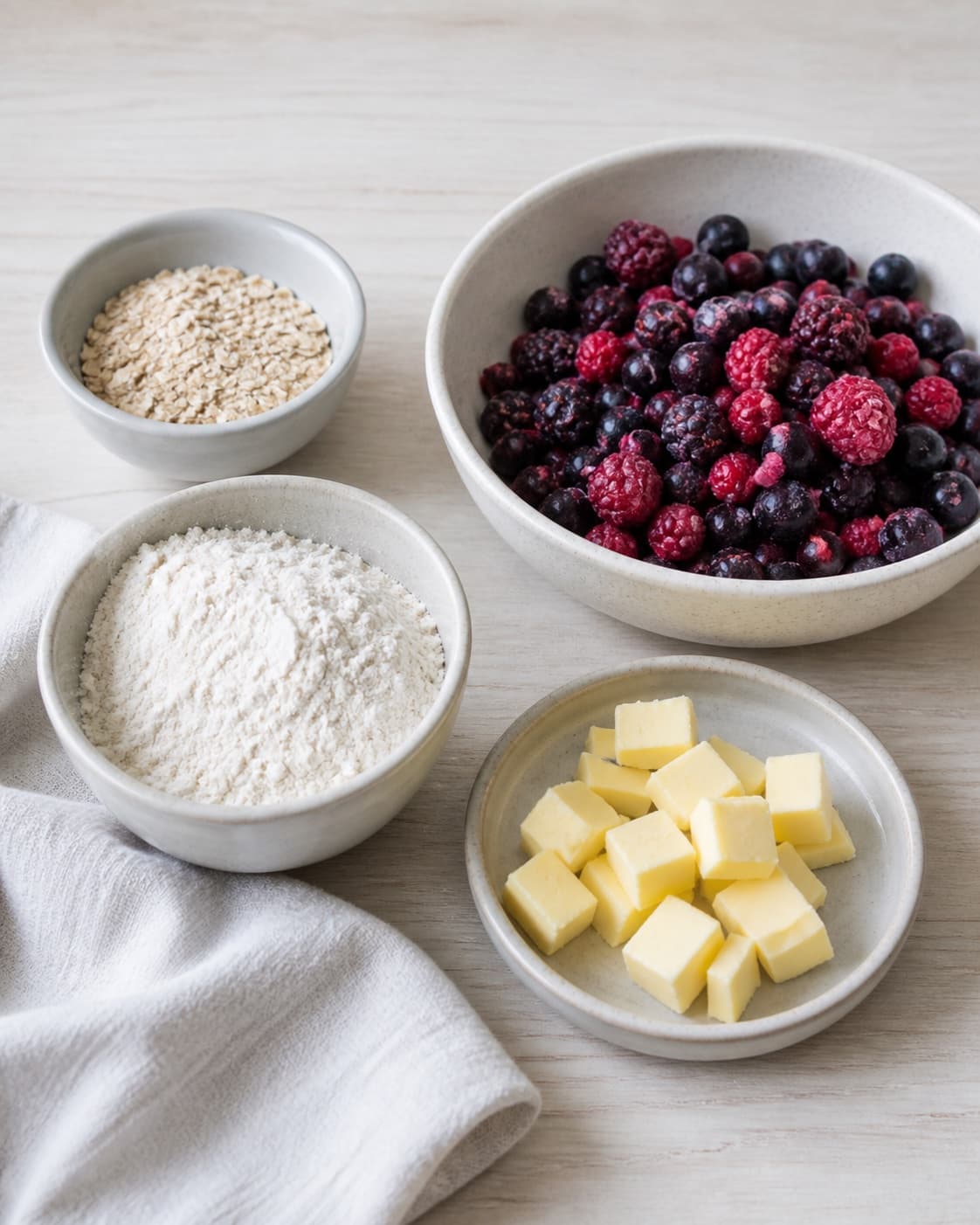 Measured ingredients for crumble med bær og havregryn til morgenmad arranged separately with blandede bær, grove havregryn, hvedemel, smør under tilberedningen af crumble med bær og havregryn til morgenmad