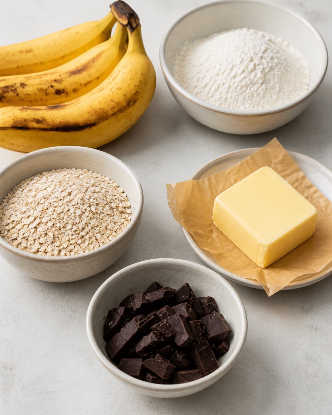 Measured ingredients for sunde bananmuffins med chokolade arranged separately with modne bananer, mørk chokolade, hvedemel, smør under tilberedningen af sunde bananmuffins med chokolade