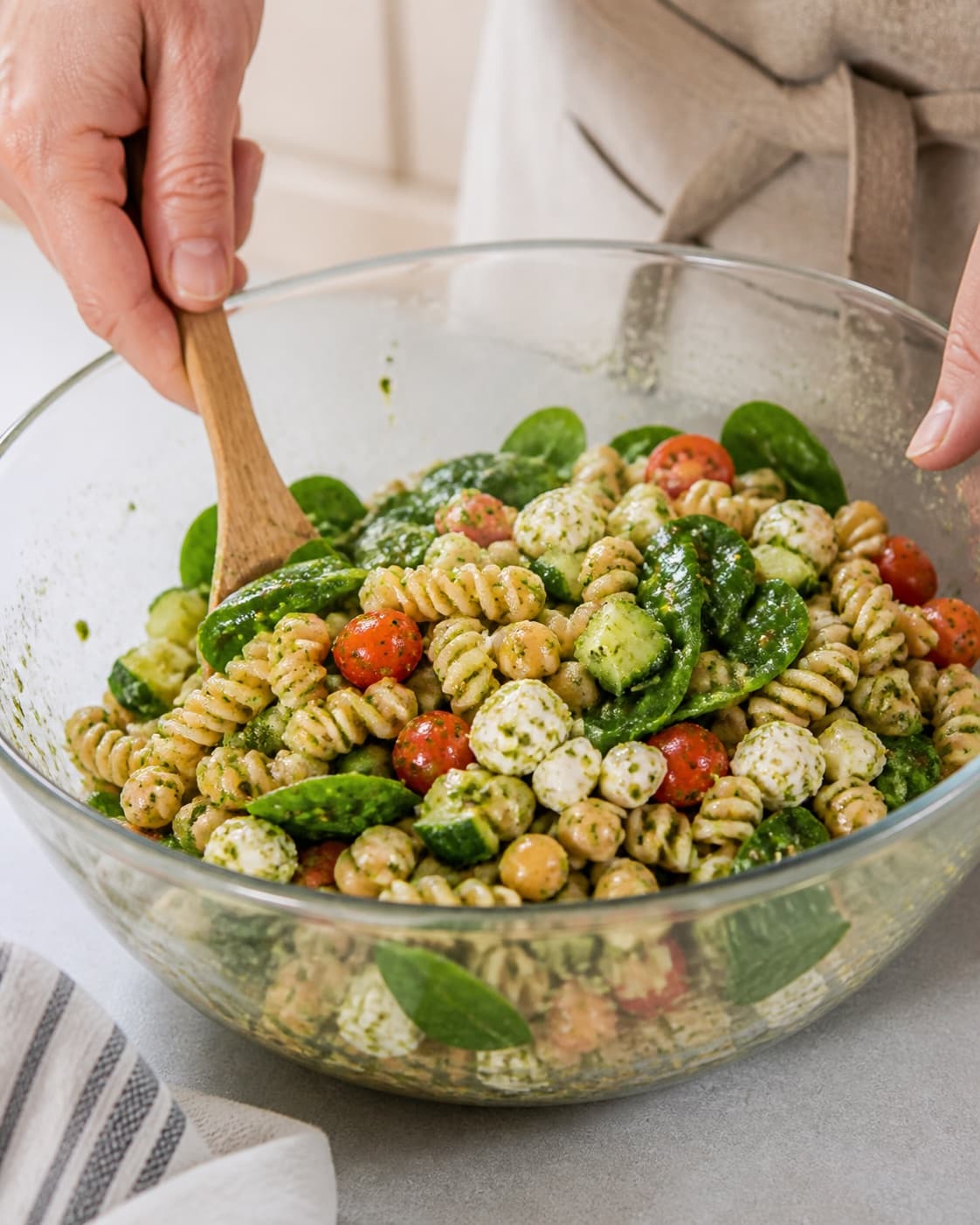 Cooled pasta chickpeas tomato cucumber spinach and feta being folded with pesto dressing under tilberedningen af pastasalat med kikærter, feta og pesto