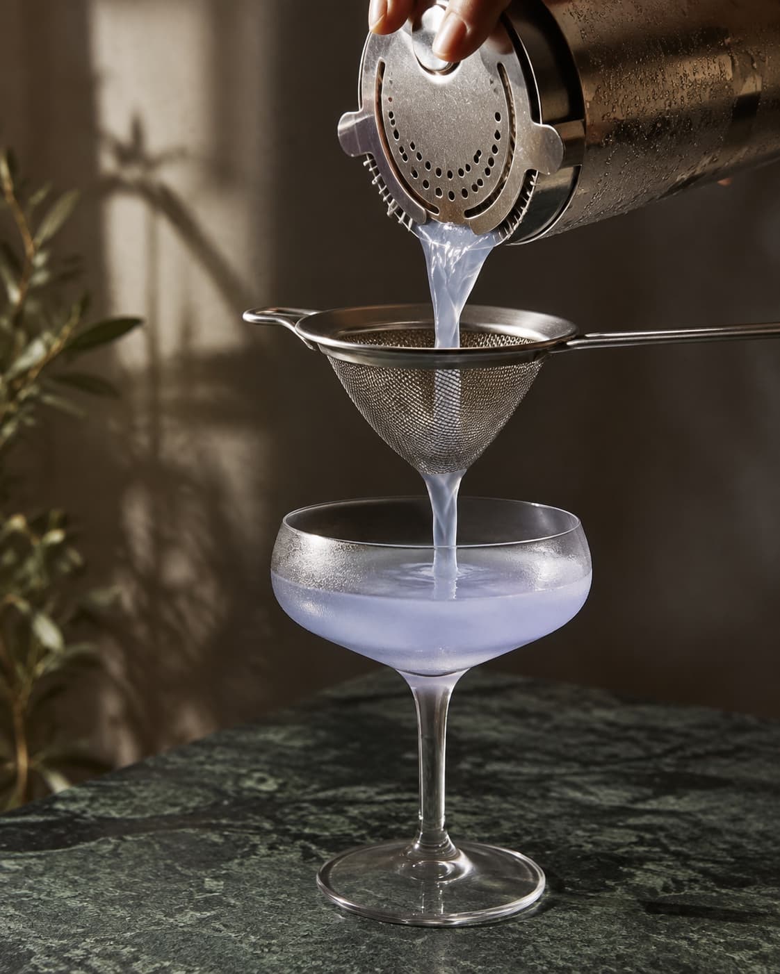 Lyseblå cocktail dobbeltsies fra shaker gennem finsigte til coupeglas