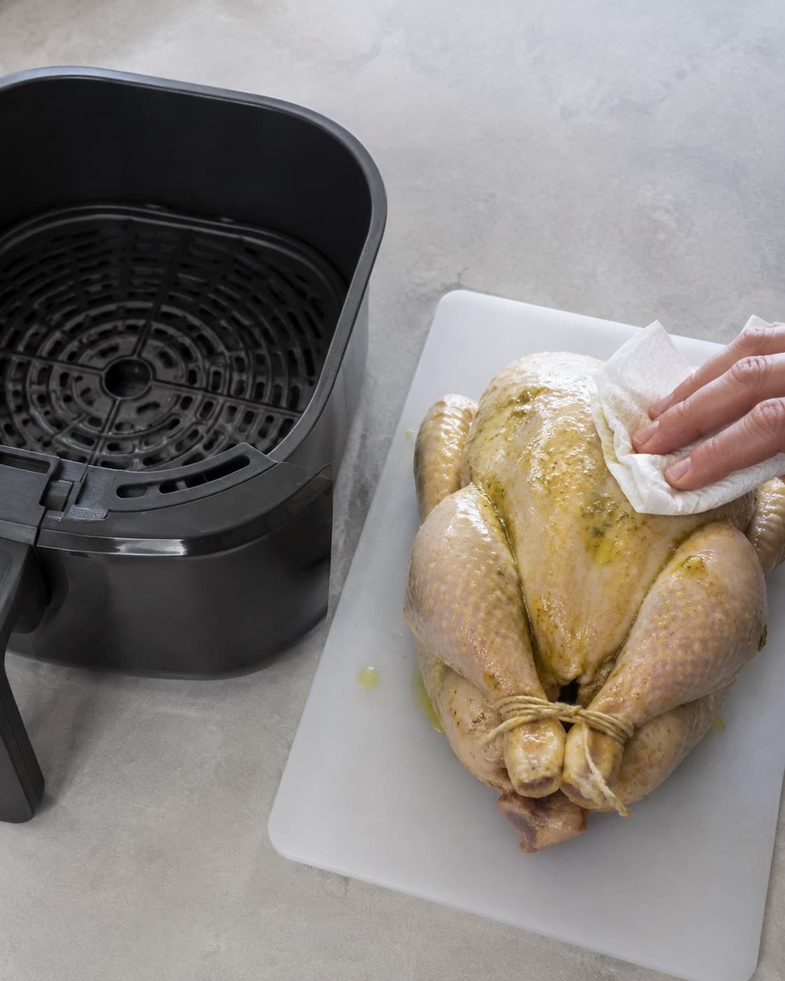 Hel rå kylling tørres og krydres på separat bræt ved siden af en tom airfryer-kurv med fri plads