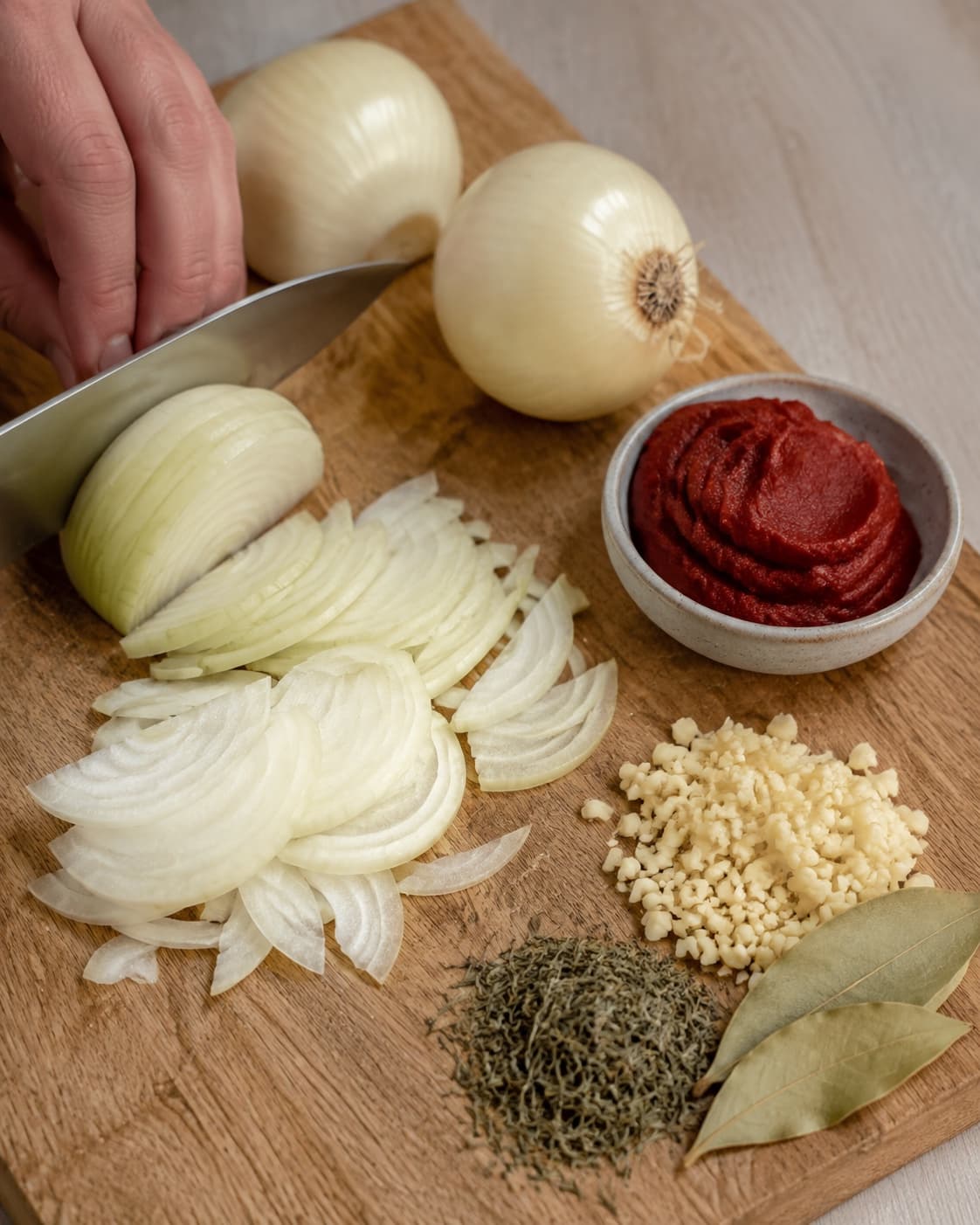 Gule løg skæres i ens tynde halvmåner ved siden af hvidløg, tomatpuré, timian og laurbær