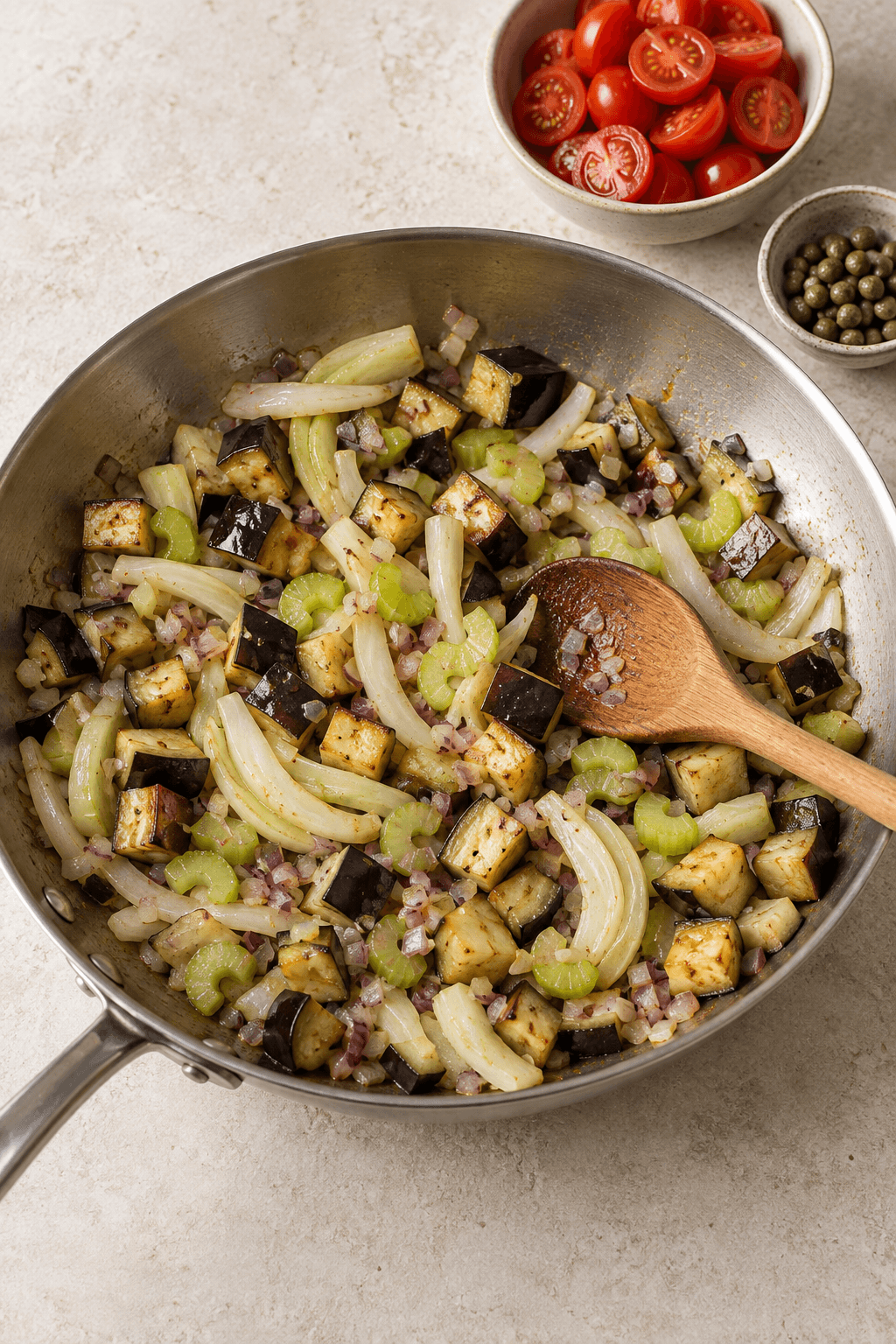 Aubergine, fennikel, selleri og rødløg brunes i én pande med tomat og kapers klar ved siden af